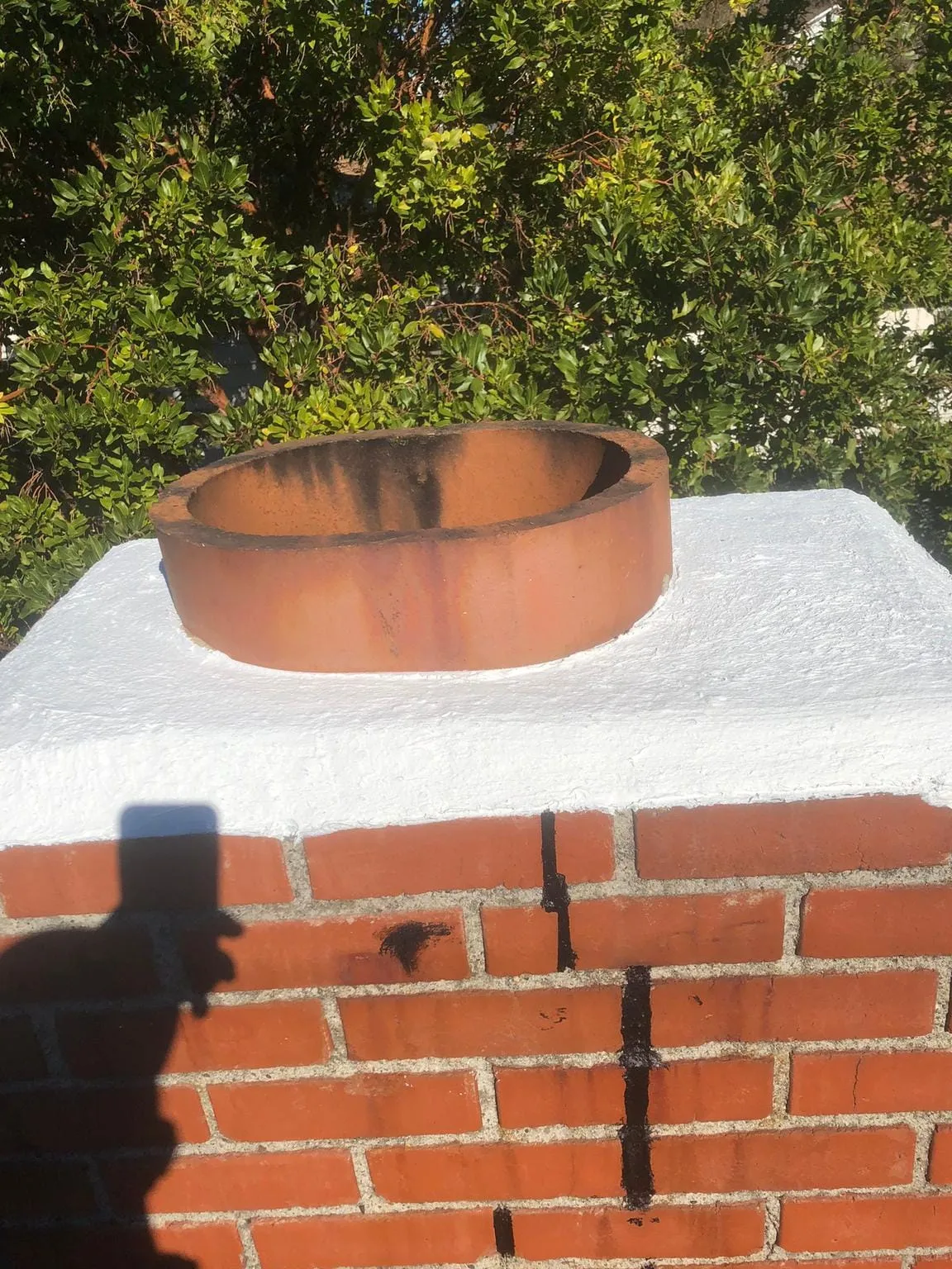 A rust-colored clay flue tile rising from a crown coated in white sealant on a red brick chimney, with dark stains running down the brick