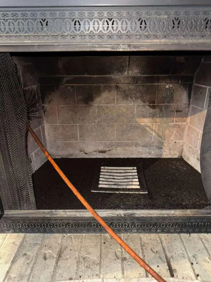 The inside of a firebox with a decorative metal frame, soot above, a cleared hearth floor with an ash-dump grate and an orange cable across it