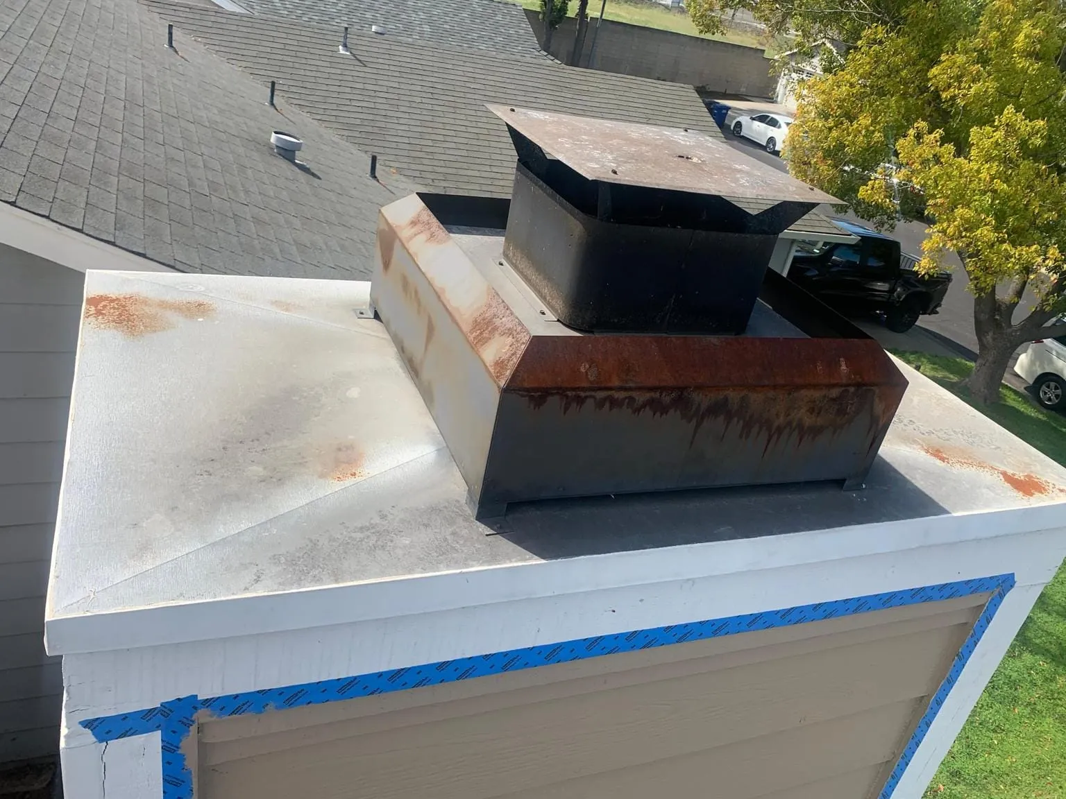A black metal cap on a rust-streaked metal chase cover, on a white-trimmed chase over tan siding, above gray shingles