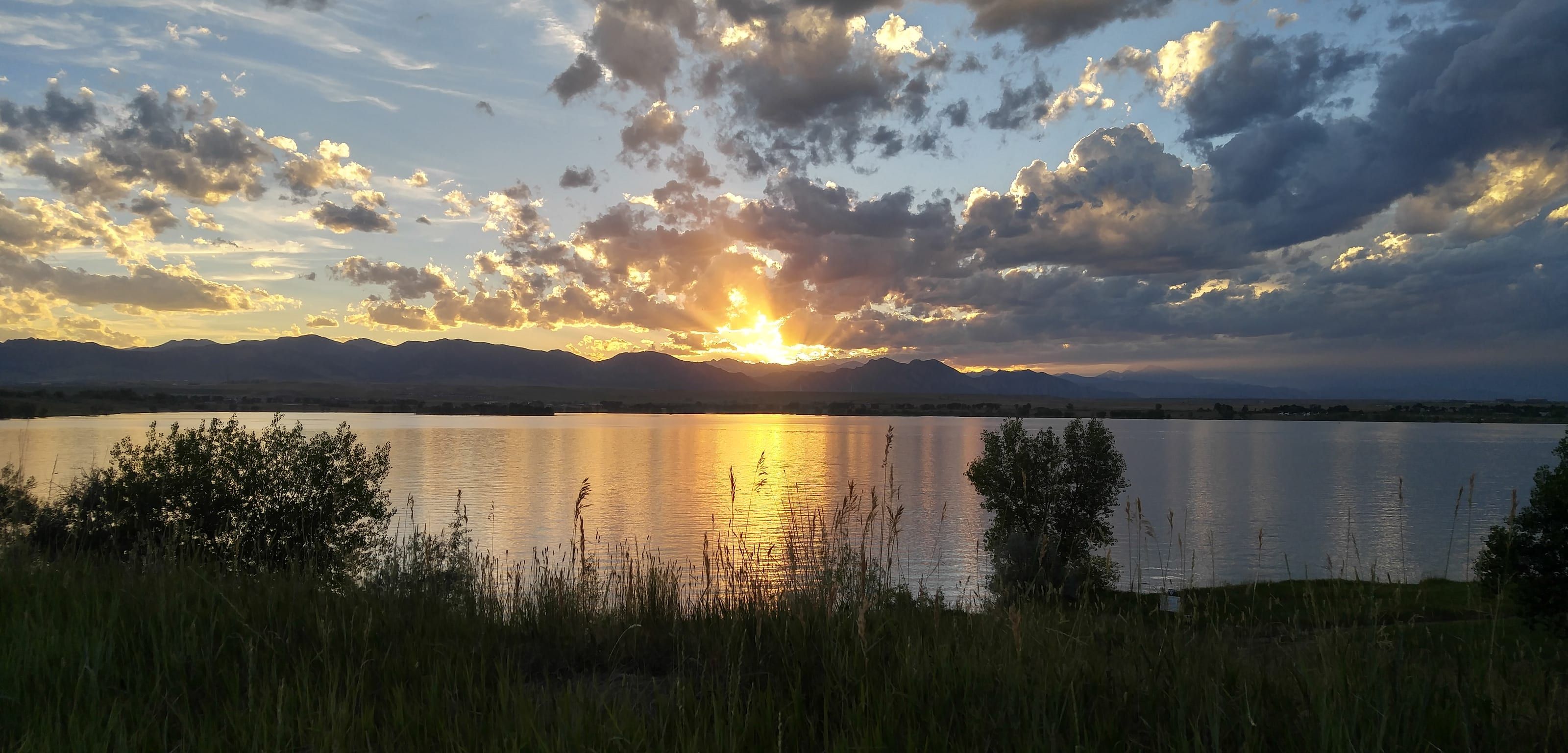 The sun going down over a wide lake, gold on the water, big clouds above and the Front Range as a dark line along the horizon